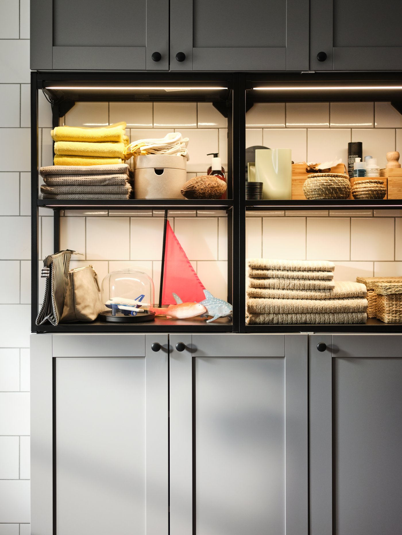 An anthracite-and-grey ENHET wall storage combination placed against a white-tiled wall and holding bathroom accessories.