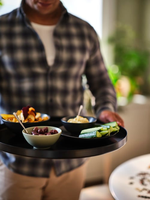 A torso of a man holding the tray section of a black GLADOM tray table, filled with bowls and plates of snacks.
