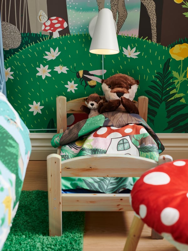 Close-up of DUKTIG doll’s bed with SKOGSDUVA soft toy otters, poster, rug and stool cover.