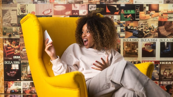Una donna seduta su una poltrona gialla STRANDMON guarda il cartellino del prezzo con aria felicemente sorpresa, la parete dietro di lei è decorata con vecchie copertine di cataloghi IKEA.