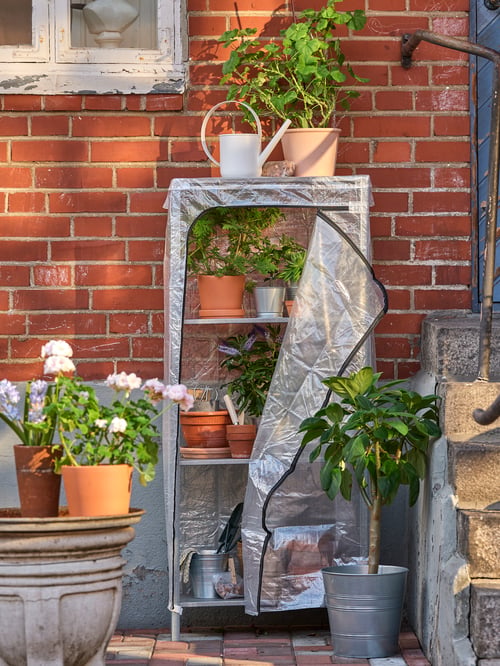 A HYLLIS shelf unit with cover set against a brick wall is used as a greenhouse for plants and storage for gardening tools.