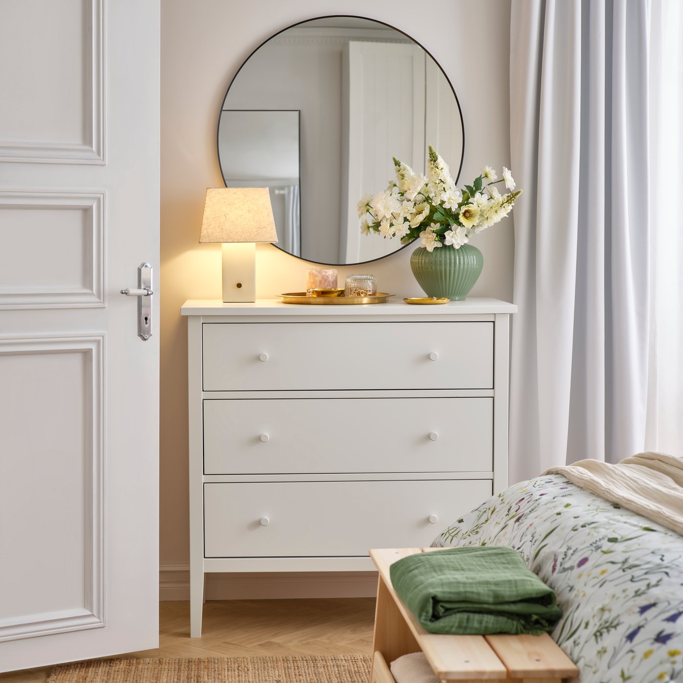 A bedroom with a round mirror over a GULLABERG chest of 3 drawers. A table lamp and flowers are on the chest of drawers.