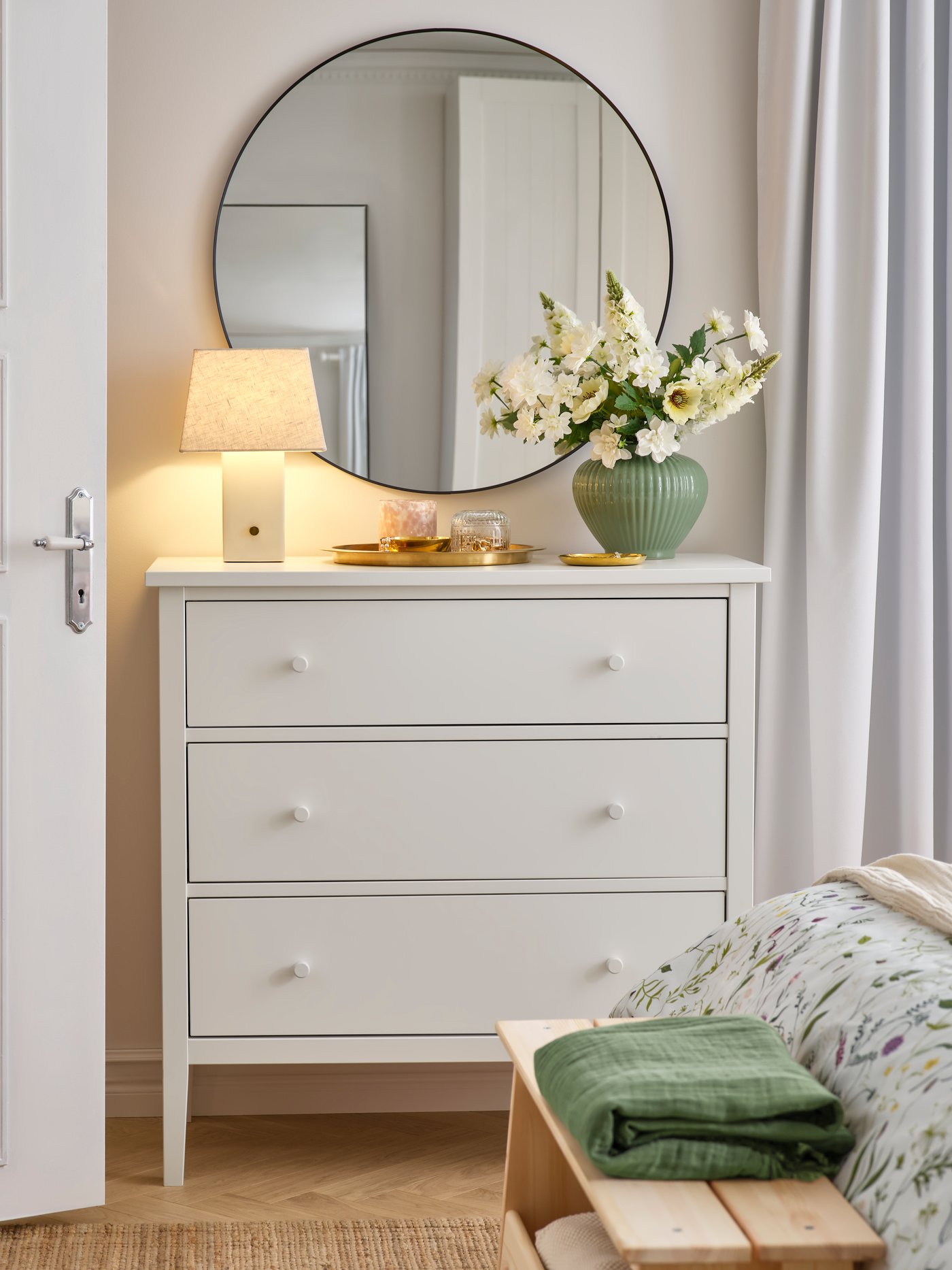 A bedroom with a round mirror over a GULLABERG chest of 3 drawers. A table lamp and flowers are on the chest of drawers.