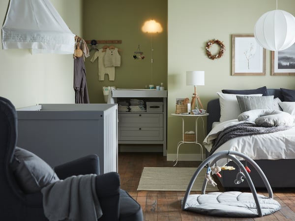 Bedroom with a grey cot, a white cradle, a dark grey divan bed, a dark blue pouffe, a round mirror and white wall lamps.