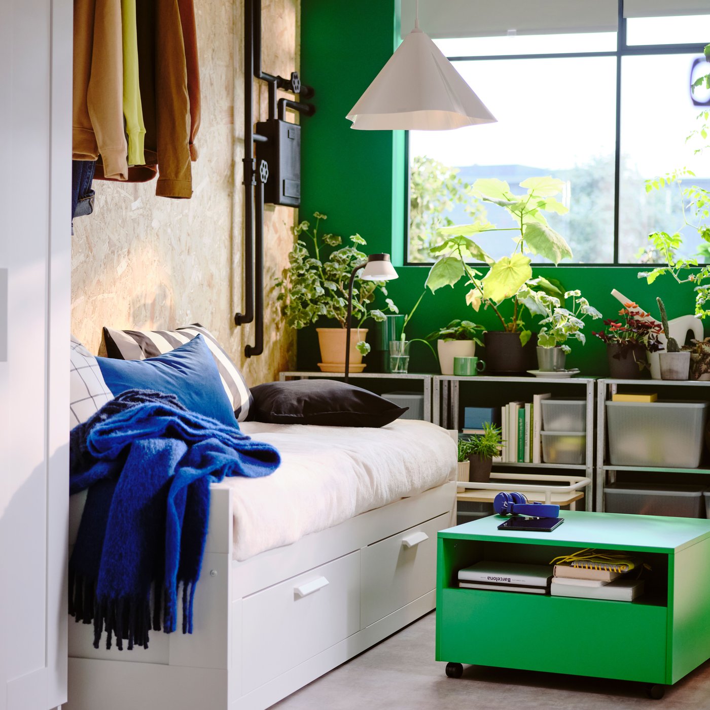 Bedroom with white daybed, a bright green table on wheels, a window and shelves with lots of green plants in the background.