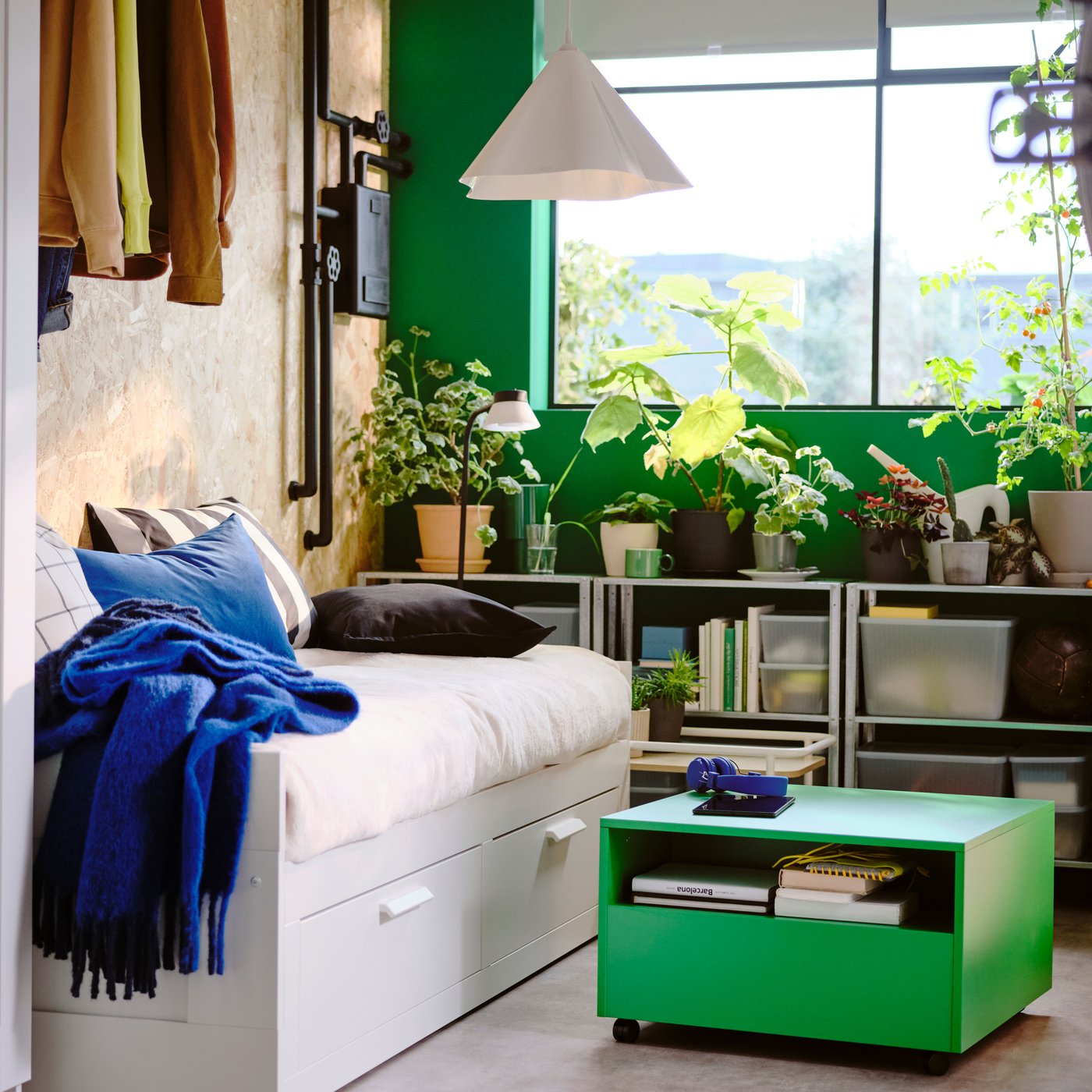 Camera da letto con letto divano bianco, un tavolino con rotelle verde vivo, una finestra e, sullo sfondo, scaffali con molte piante verdi.
