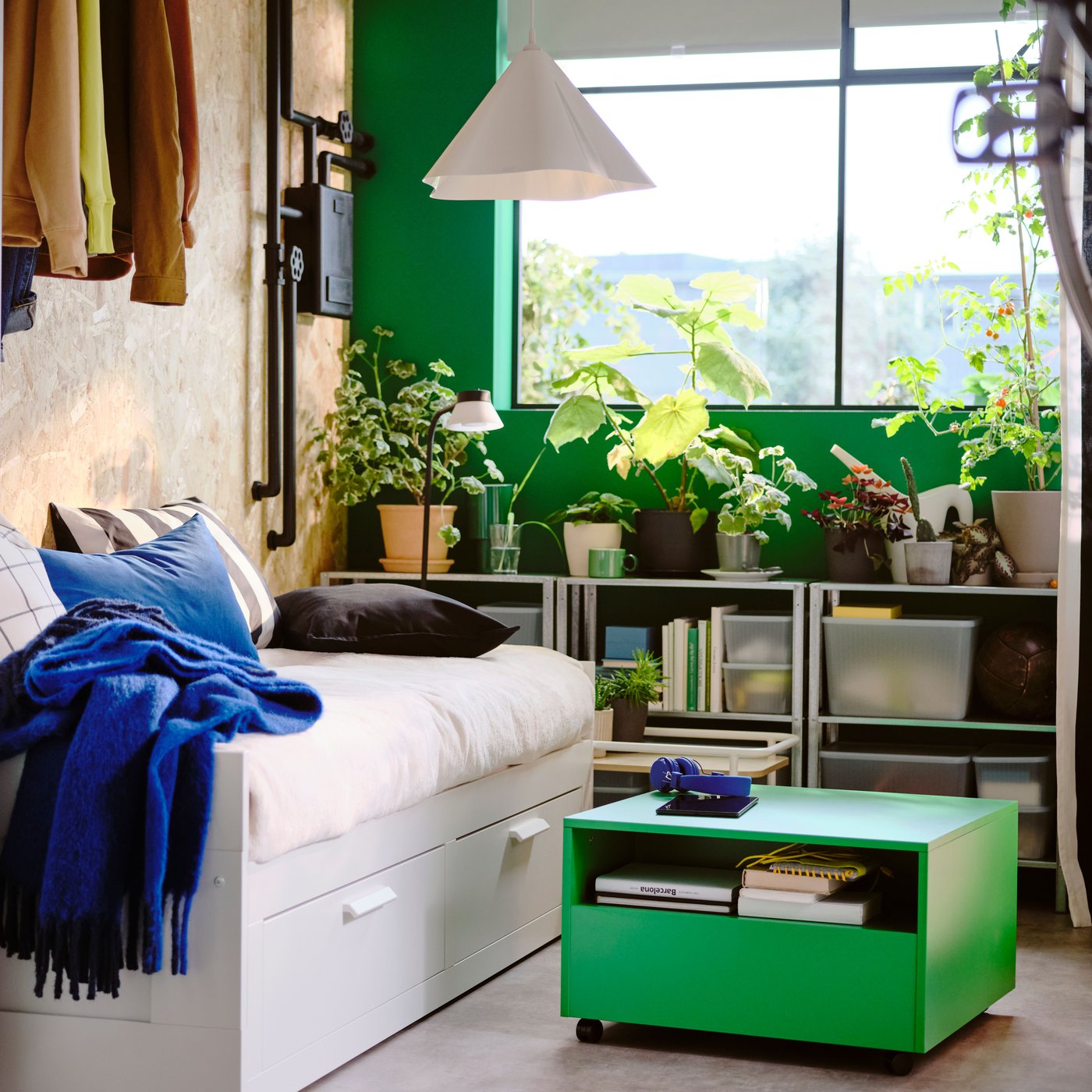 Es ist ein Schlafzimmer mit weißem Tagesbett, einem leuchtend grünen Tisch auf Rollen, einem Fenster und Regalen mit vielen Grünpflanzen zu sehen.