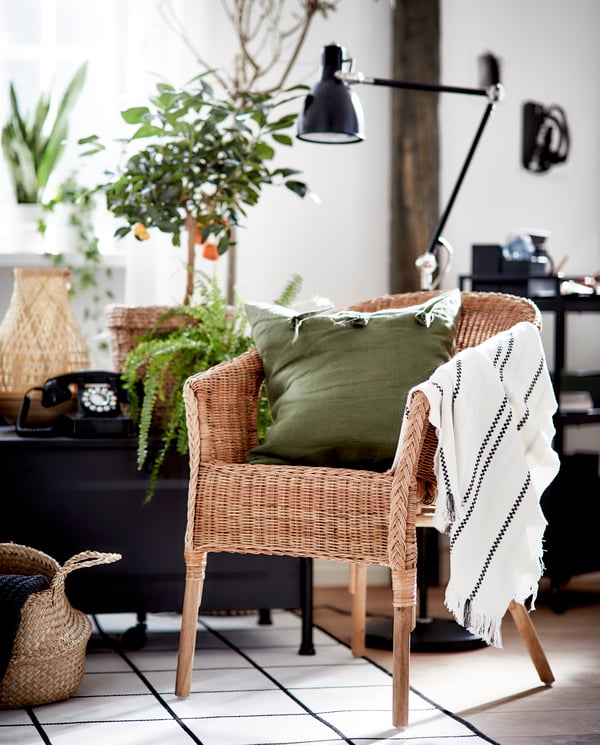 Poltrona in rattan AGEN con cuscino beige NORNA, plaid verde scuro, cuscini con federe AINA, tazza di tè su un tavolino e tappeto a tessitura piatta LOHALS.