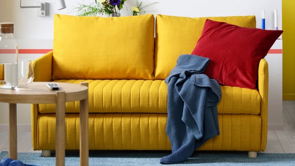 Gul FRIDHULT bäddsoffa med ribbad front och färgglada kuddar i ett ljust vardagsrum.