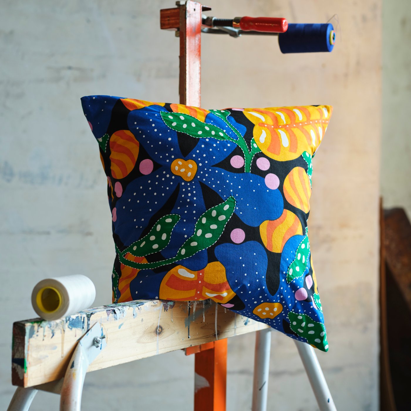 A MÄVINN cushion cover with a multicoloured floral pattern rests on a wooden beam stained with old paint.