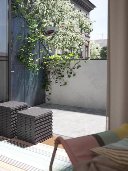 Een modern balkon is voorzien van grijze IKEA RUNNEN vlonders, met weelderige groene klimop die tegen een donkere muur omhoog kruipt, gezien vanuit een binnenruimte met een kleurrijke stoel en houten vloer.