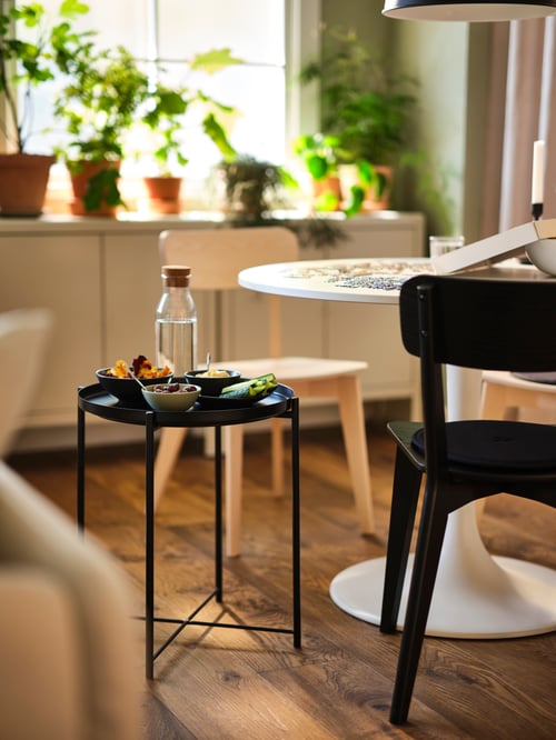 A black GLADOM tray table covered in snacks next to a white DOCKSTA table with two LISABO chairs in black and ash.