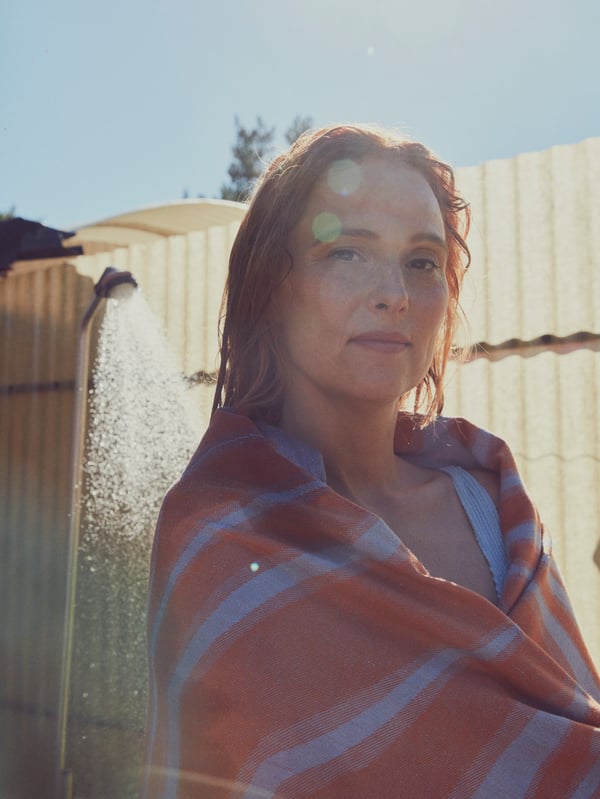 A woman smiles and stands close to an outdoor shower, with a blue and orange BASTUA bath sheet wrapped around her shoulders.