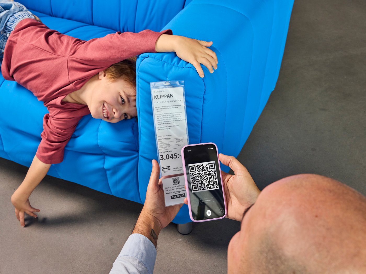 Dziecko wygłupiające się na niebieskiej sofie IKEA, obok taty, który skanuje price tag