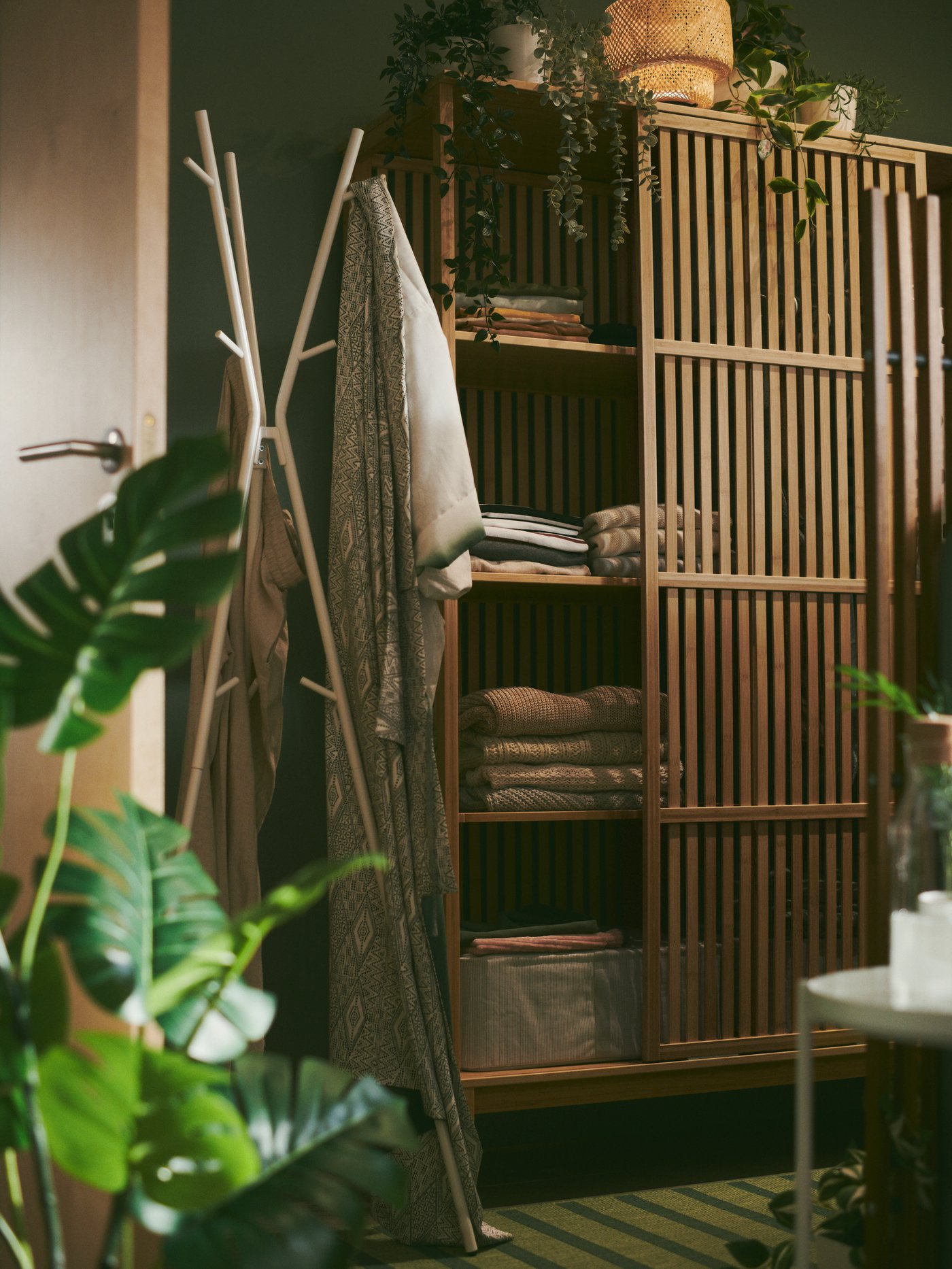 Ein offener NORDKISA Kleiderschrank mit Schiebetür aus Bambus ist in einem grünen Schlafzimmer zu sehen. Auf den Böden liegt gefaltete Kleidung.