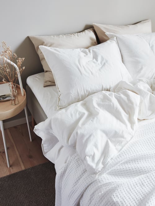 Chambre à coucher avec une housse de couette blanche avec 2 taies d’oreiller en coton, 2 taies d’oreiller beiges et un couvre-lit à côté d’une table d’appoint.