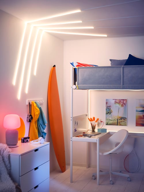 Dans une chambre, un lit mezzanine VITVAL, une planche de surf, des baguettes lumineuses à DEL MYRVARV fixées au mur et une lampe de table avec haut-parleur wifi.