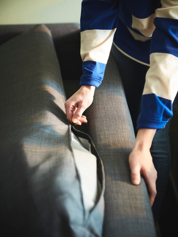 A person stands by a dark-grey FRIHETEN sofa-bed, pulling on the partly open zipper of a KLAGSHAMN back-cushion cover.