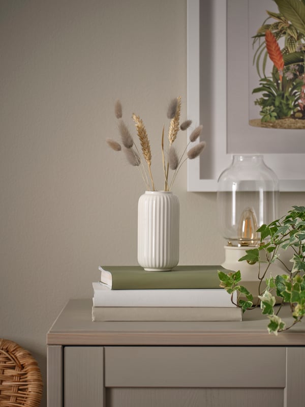 Eine kleine, beige, geriffelte STILREN Vase steht auf einem Stapel Bücher auf einem Sideboard, daneben eine Tischleuchte, darin ein paar Büschel Hasenschwanzgras.