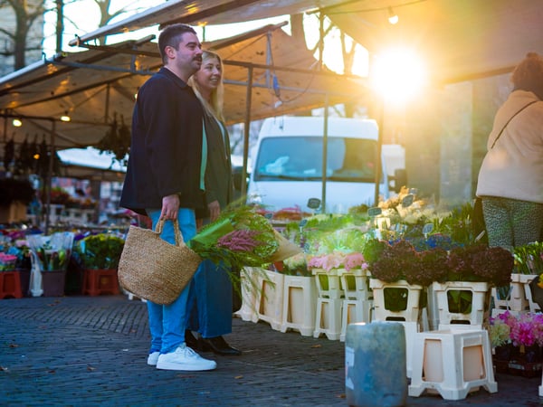 A man and a woman standing by a florist stand at an outdoor market, carrying a woven basket filled with fresh flowers.