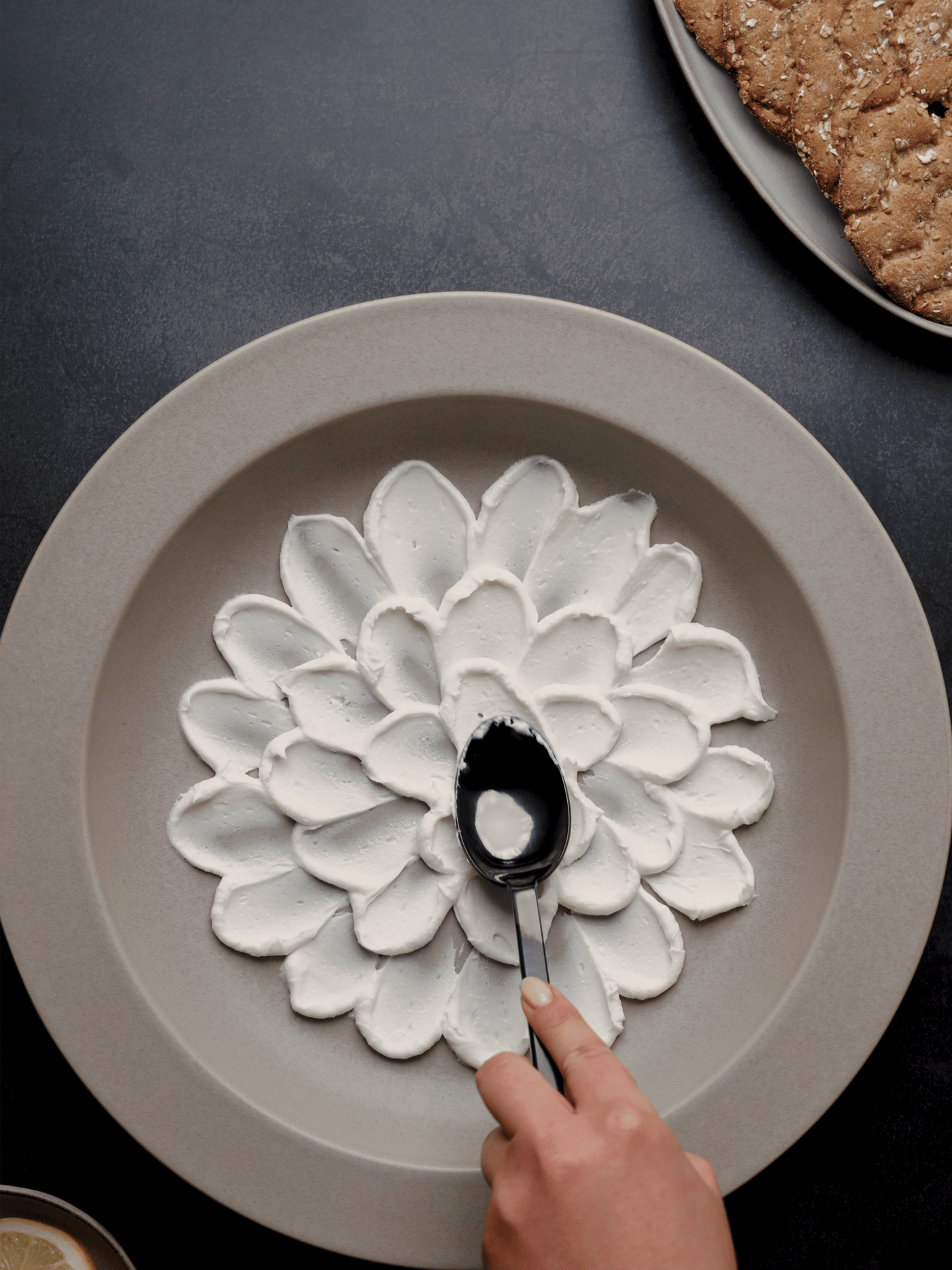 Eine Hand, die Schlagsahne in einem Blumenmuster auf einem grossen Teller verteilt, mit einem Backwerk im Hintergrund.
