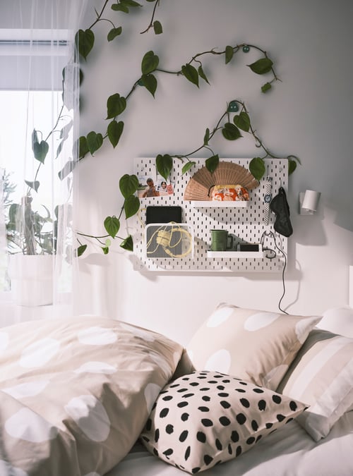 Bedroom with a white pegboard made of fibreboard supporting 2 shelves next to a wall reading lamp above a cushion cover.