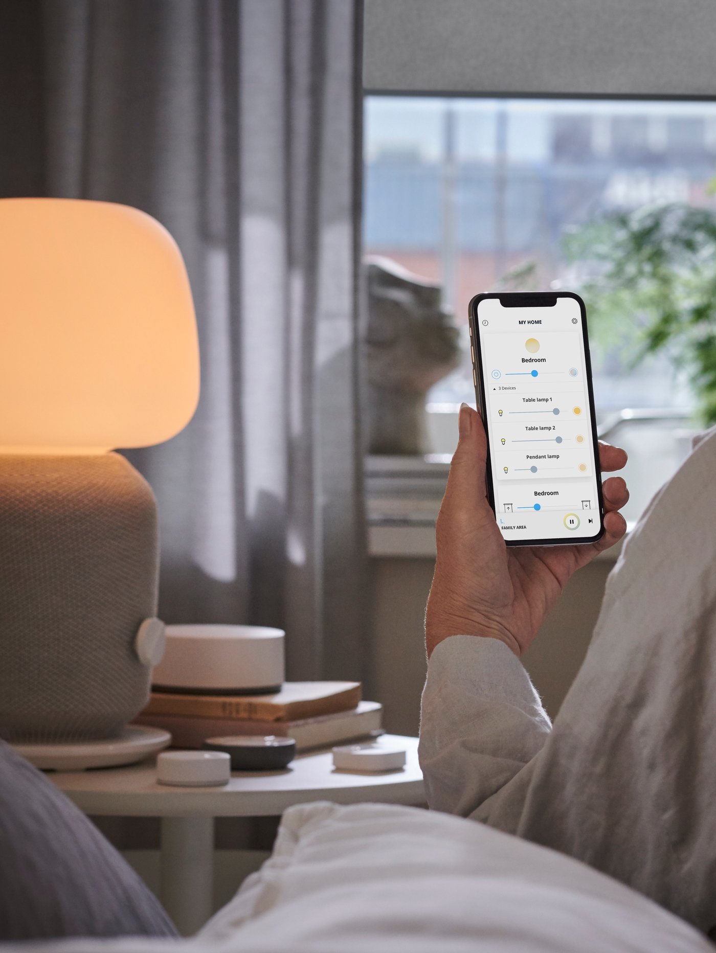A hand holding a mobile device in front of a table with a speaker lamp and remote controls, window in the background.