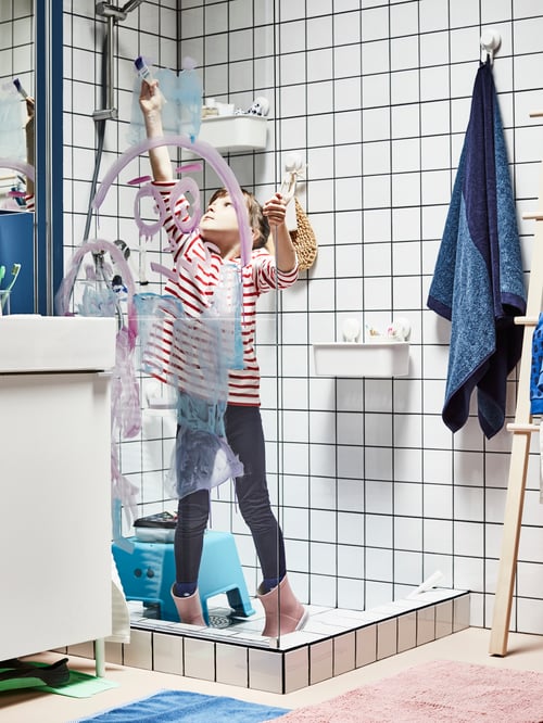 Un enfant dans une salle de bains. Il y a une douche et un panier en plastique blanc muni de ventouses. Il contient divers accessoires. 