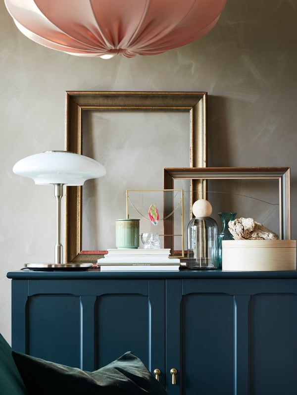 Cornici dorate, libri, oggetti decorativi e una lampada da tavolo in vetro nichelato/bianco opalino su un mobile blu-verde.