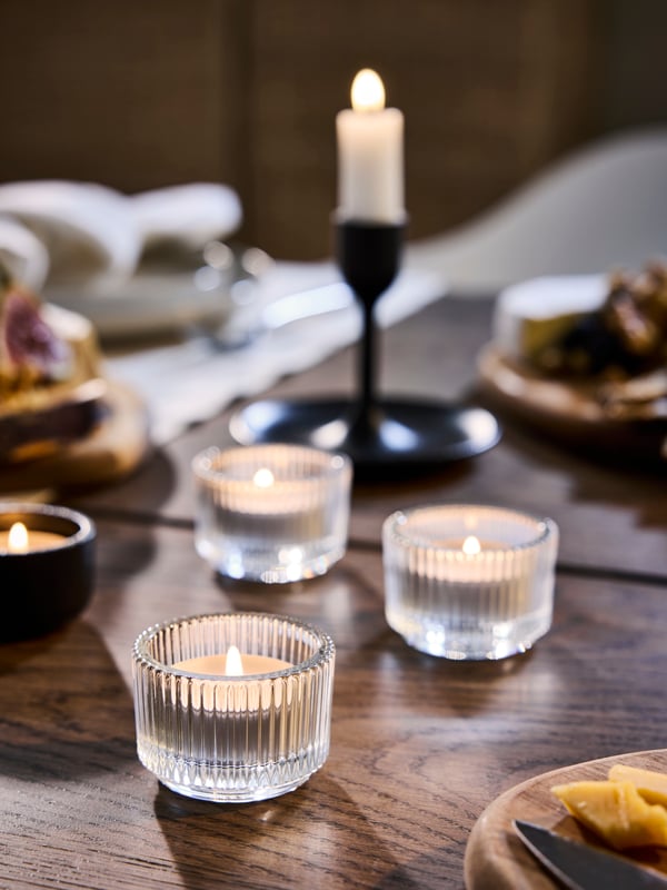 Three glass FINSMAK tealights sitting on a dark wood table with a black candle stick holder in the background.