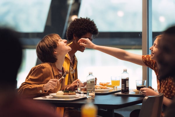 Trei persoane stau la masă într-un restaurant, mâncând și râzând; una hrănește pe alta cu un cartof prăjit. Pe masă sunt farfurii cu mâncare, pahare și sticle de apă.