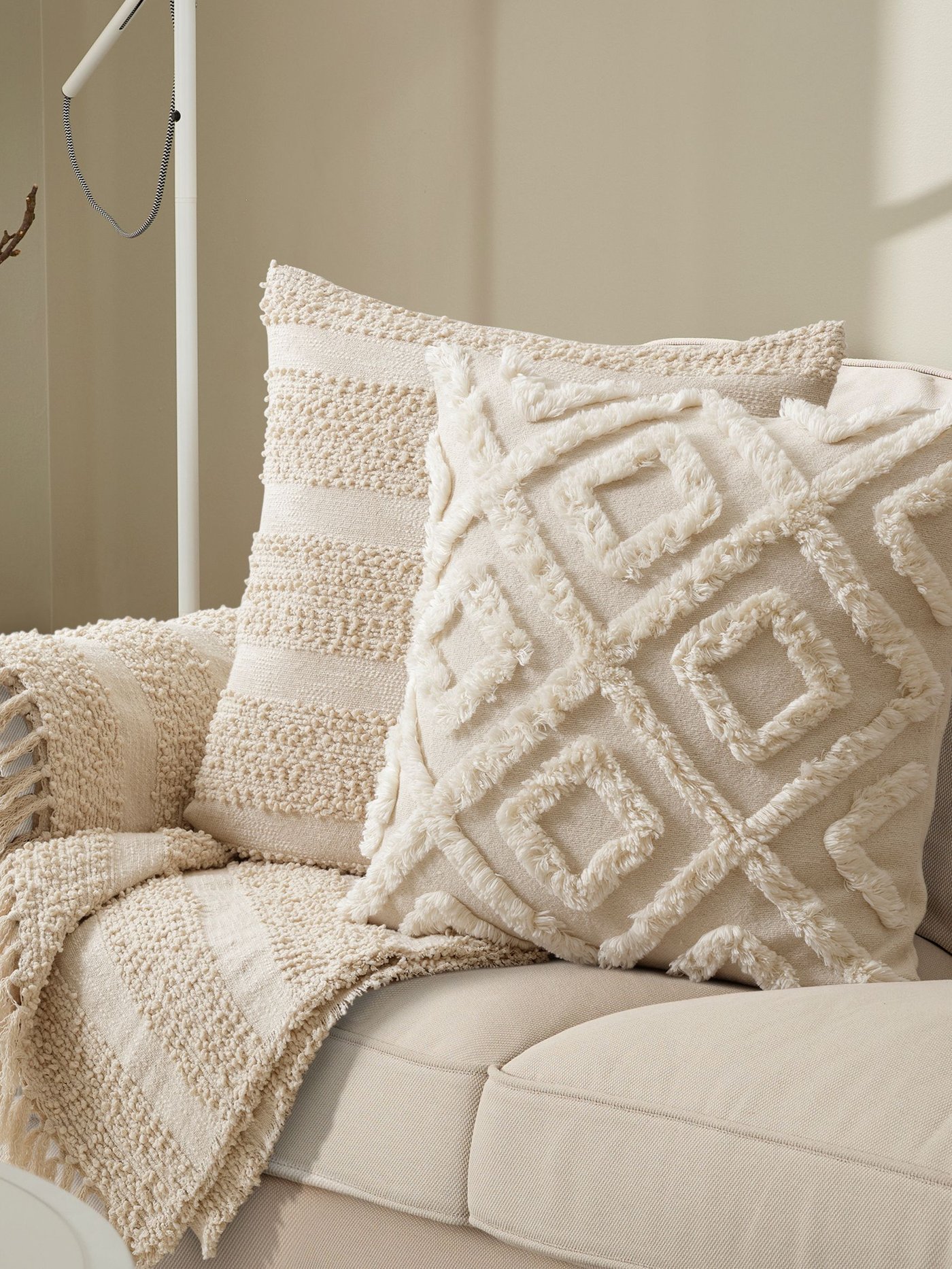 Auf einem beigen Sofa liegt ein naturfarbenes EKGULDMAL Kissen mit geometrischem Muster im Boho-Stil und ein naturfarbenes GÖLSTARR Kissen mit Struktur. Über der Armlehne des Sofas liegt ein passendes GÖLSTARR Plaid in der gleichen Farbe.