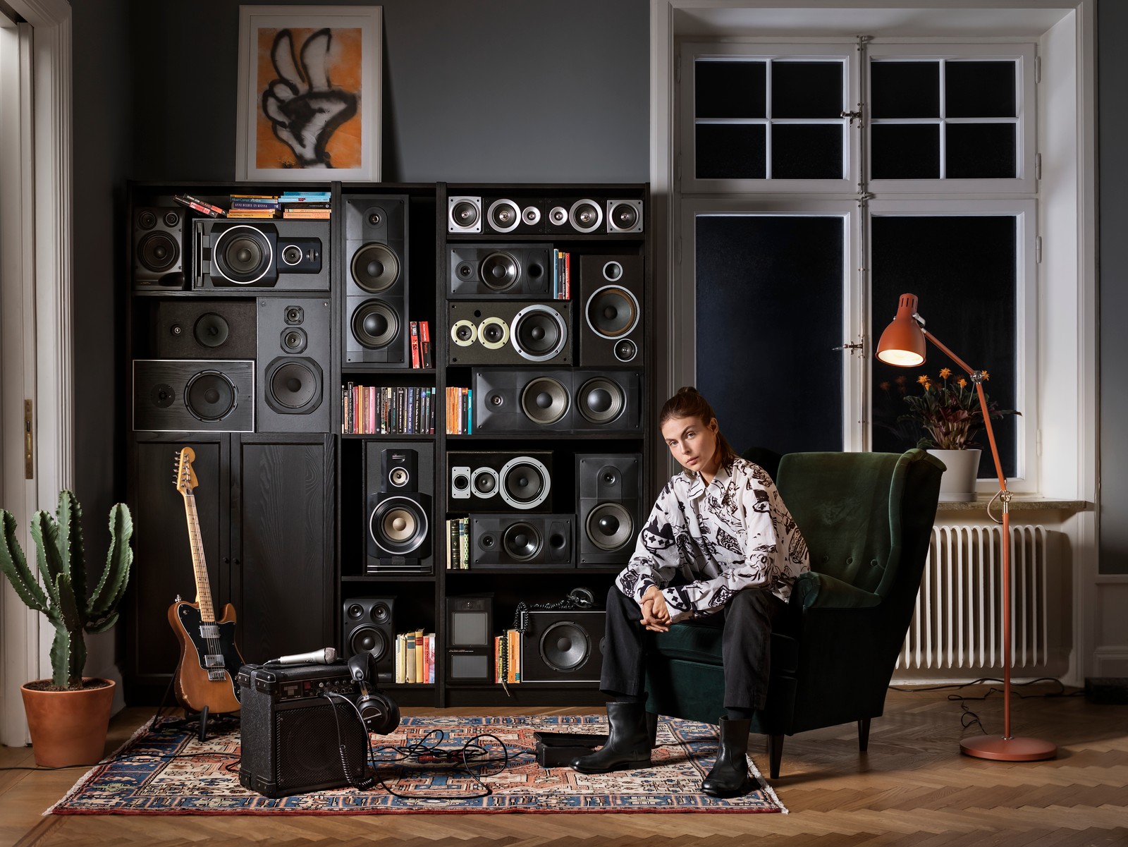 Mlada žena sedi na STRANDMON fotelji. Na podu pored nje nalaze se električna gitara i pojačalo. Na zidu iza nje nalazi se velika polica za knjige puna zvučnika.