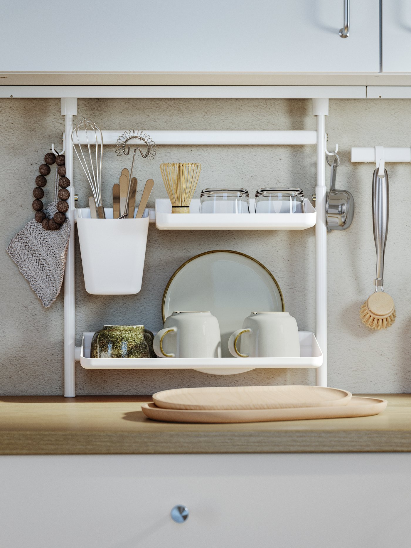 White kitchen wall rack neatly organised with utensils in a hanging container, glass jars, mugs, and a plate, alongside a whisk, wooden brush, and textiles, placed above a wooden countertop.