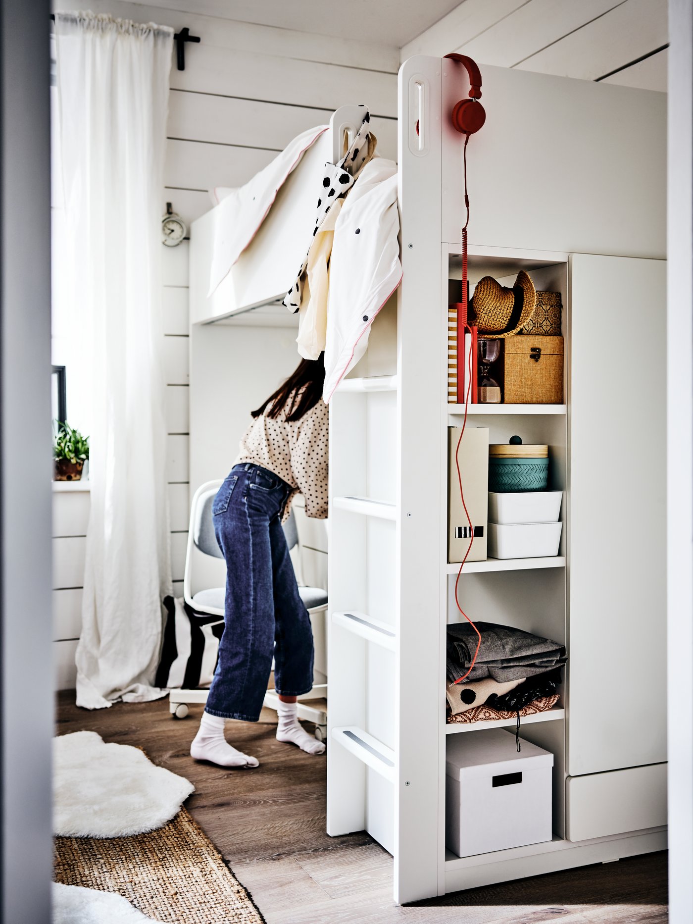 A girl standing by an ÖRFJÄLL chair at her desk under a white loft bed. Storage boxes are on shelves in the bed frame.