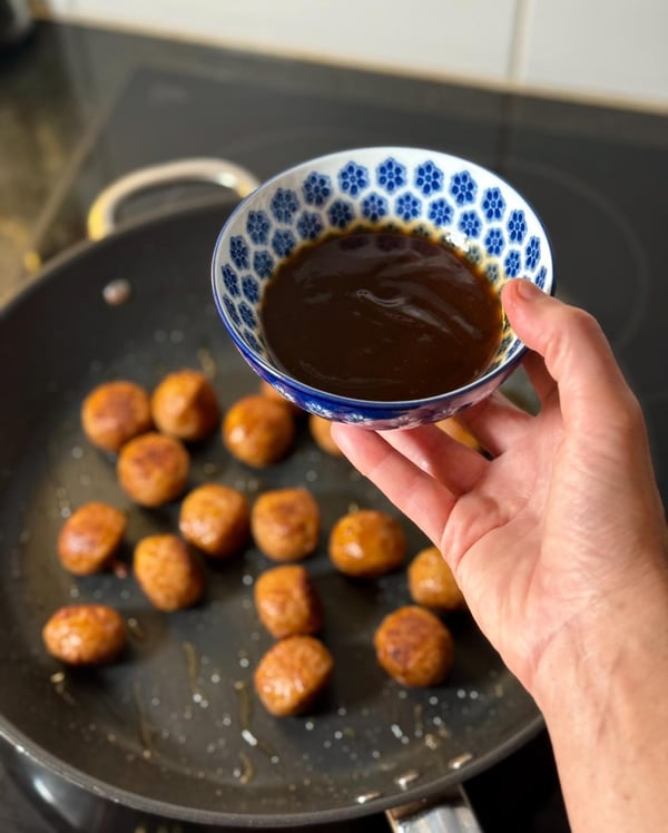One hand holds a bowl of sauce over a pan full of golden brown meatballs cooking on the stove.