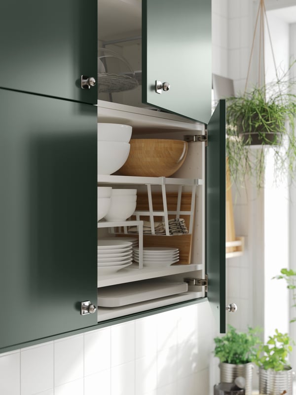 A wall cabinet with deep green kitchen fronts and doors ajar showing plates, bowls, a shelf insert and a clip-on basket.
