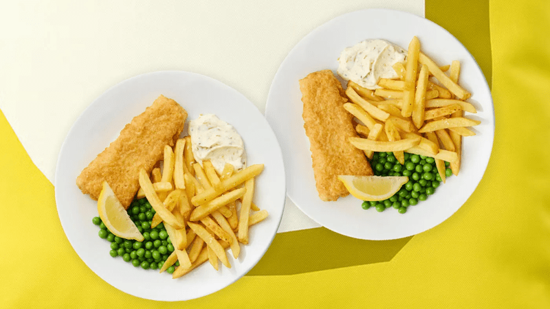 Värikästä taustaa vasten on kaksi Fish & chips -annosta, joissa kummeliturskafile, ranskalaisia, herneitä, majoneesi ja sitruunan pala.