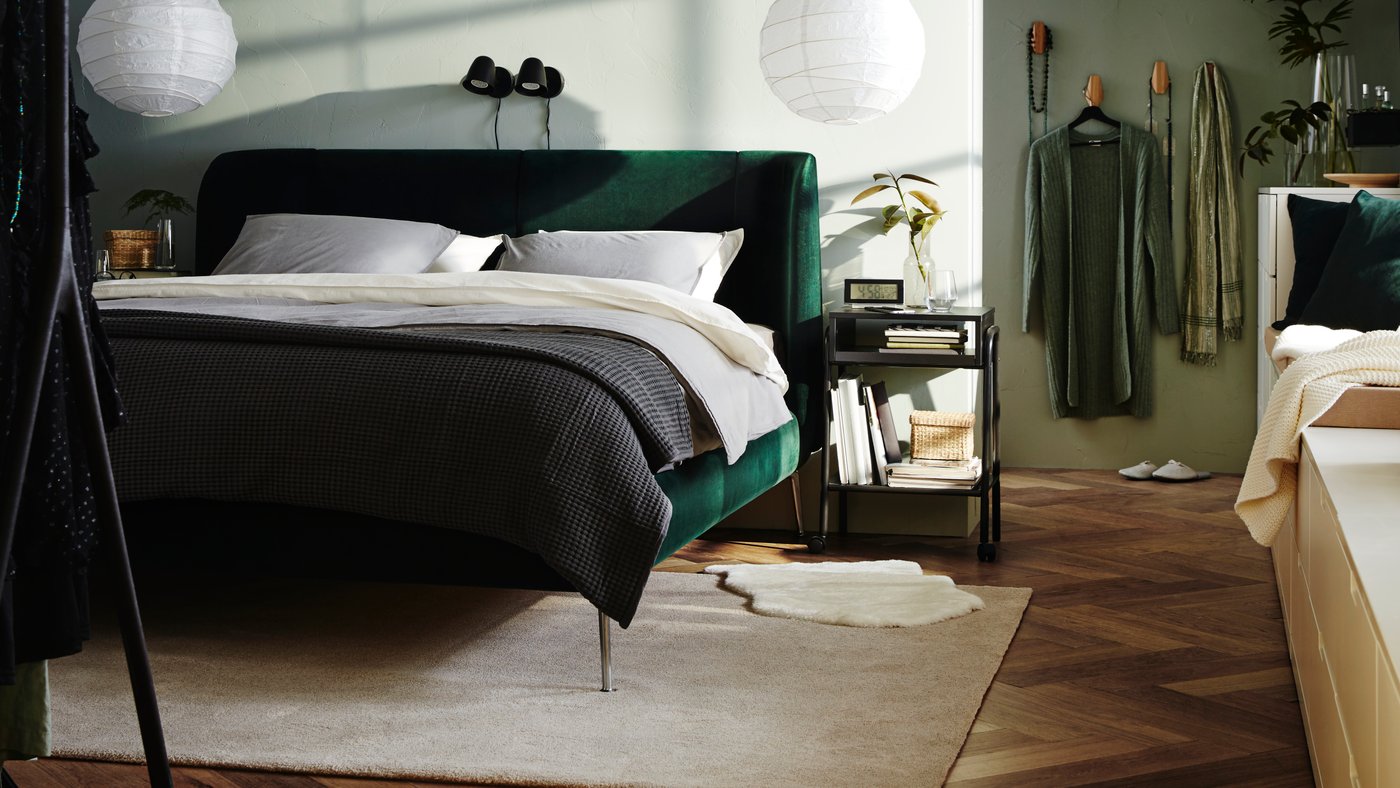 A bedroom with a TUFJORD dark green upholstered bed frame, 2 white pendant lamp shades, white bedding and a grey bedspread.