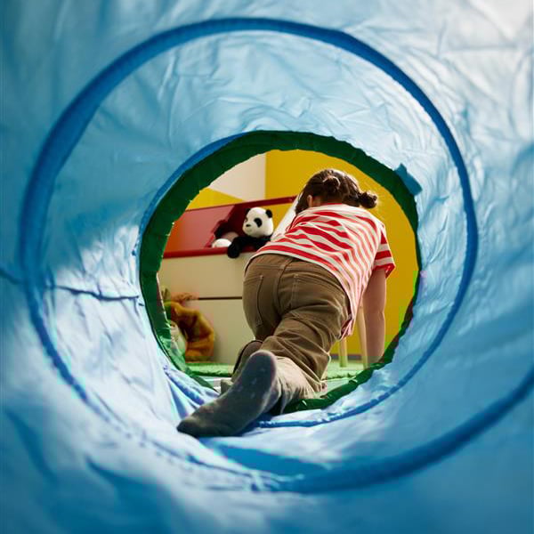 Bambina che gioca e attraversa un tunnel morbido blu in una cameretta colorata, con peluche appoggiati su uno scaffale sullo sfondo.