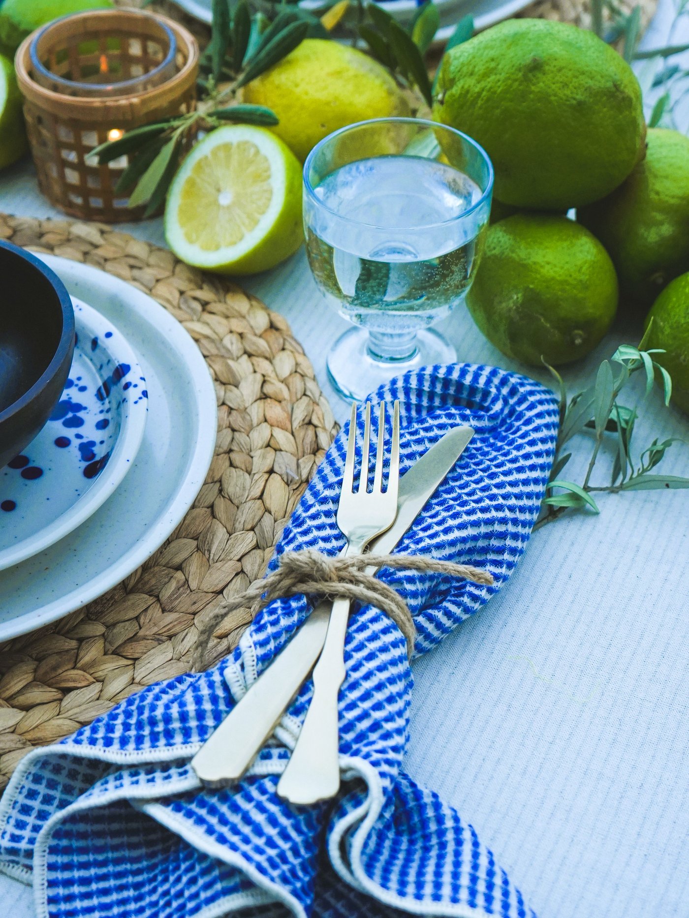 Une table dressée avec de la vaisselle SILVERSIDA bleue et blanche, des couverts posés sur une serviette VÄLVÅRDAD à carreaux, des citrons frais, des citrons verts et des brins d'olivier pour créer une ambiance estivale.