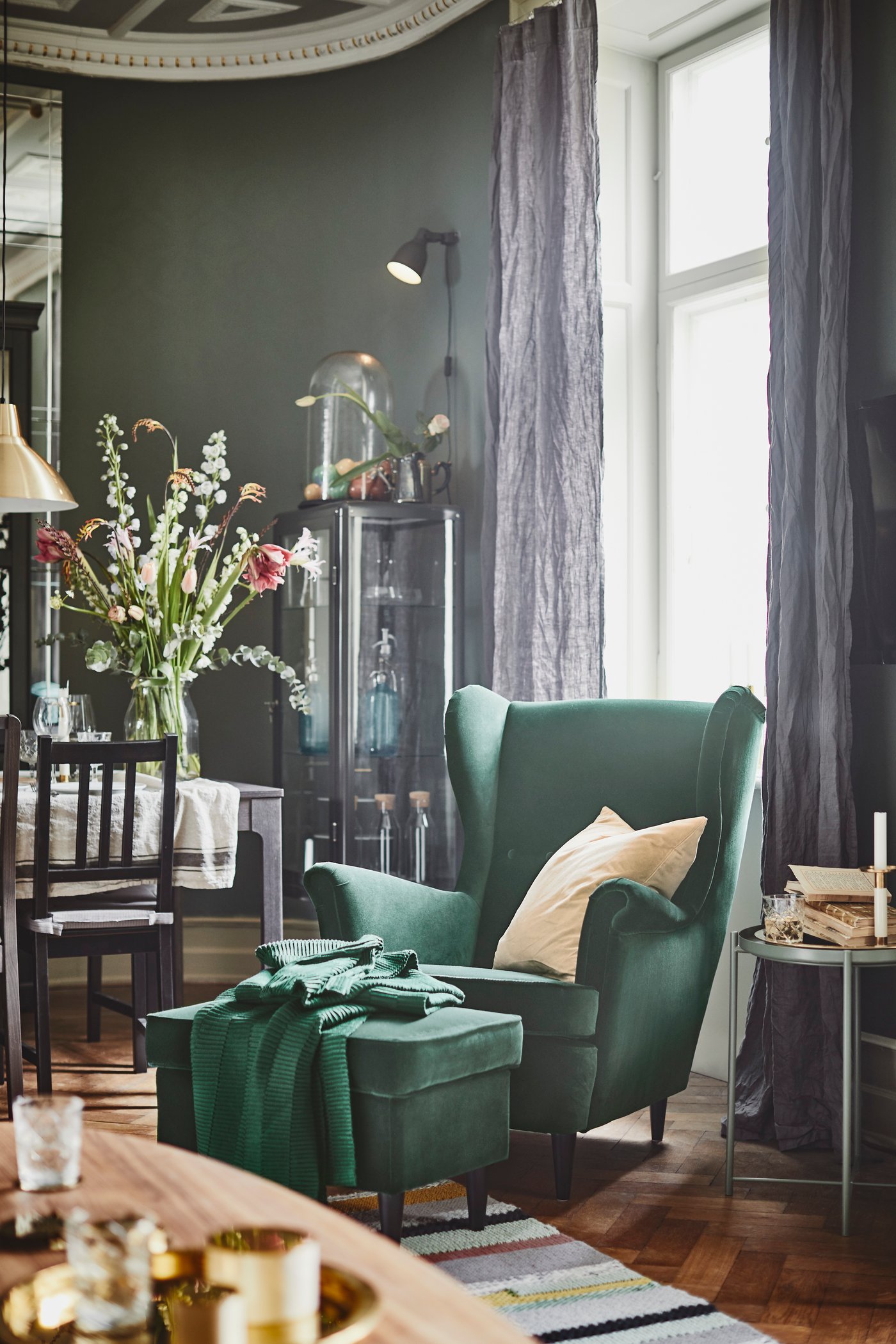 A wing chair in green velvet with a matching stool stands in front of a window with dark grey linen curtains and next to a dining table on which a large vase with flowers stands
