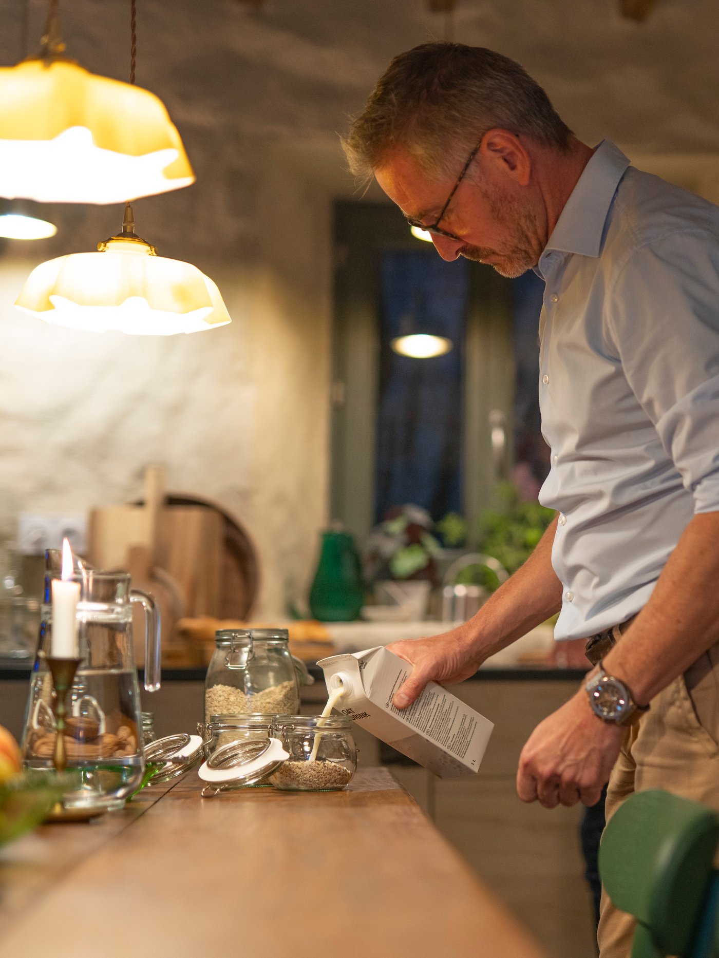 Un homme dans la cuisine le soir. Il verse du lait d’avoine dans un bocal KORKEN rempli de flocons d’avoine.