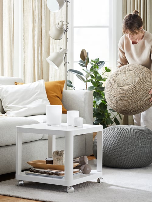 A white TINGBY side table on castors holds candles and dumbbells by a sofa. A person is stacking woven pouffes nearby.