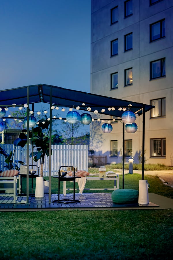 GUNNÖN pavillon danner rammen om et festligt, pyntet udendørsområde med SOLVINDEN solcellelamper, havemøbler, grønne planter og gulvbelægning.