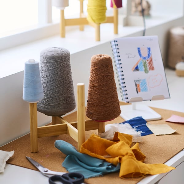 A bamboo DAKSJUS plant stand on top of a white desk holding different coloured rolls of sewing twine, with scissors nearby.