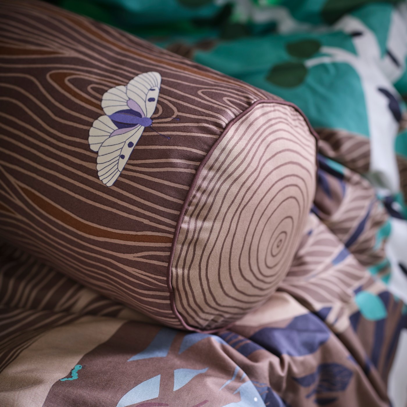 A SKOGSDUVA cylinder-shaped cushion is placed on a SKOGSDUVA duvet cover and pillowcase with a forest pattern.