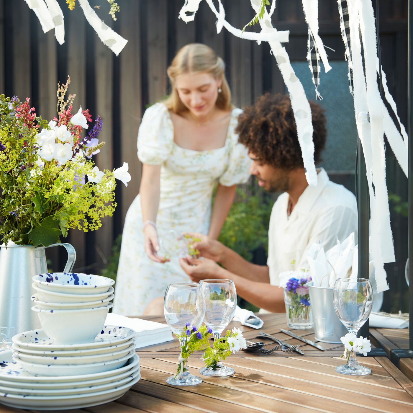 Ein Mann sitzt an einem Esstisch im Freien, während eine Frau neben ihm steht. Beide arbeiten gemeinsam an der Dekoration für das Midsommar-Fest.