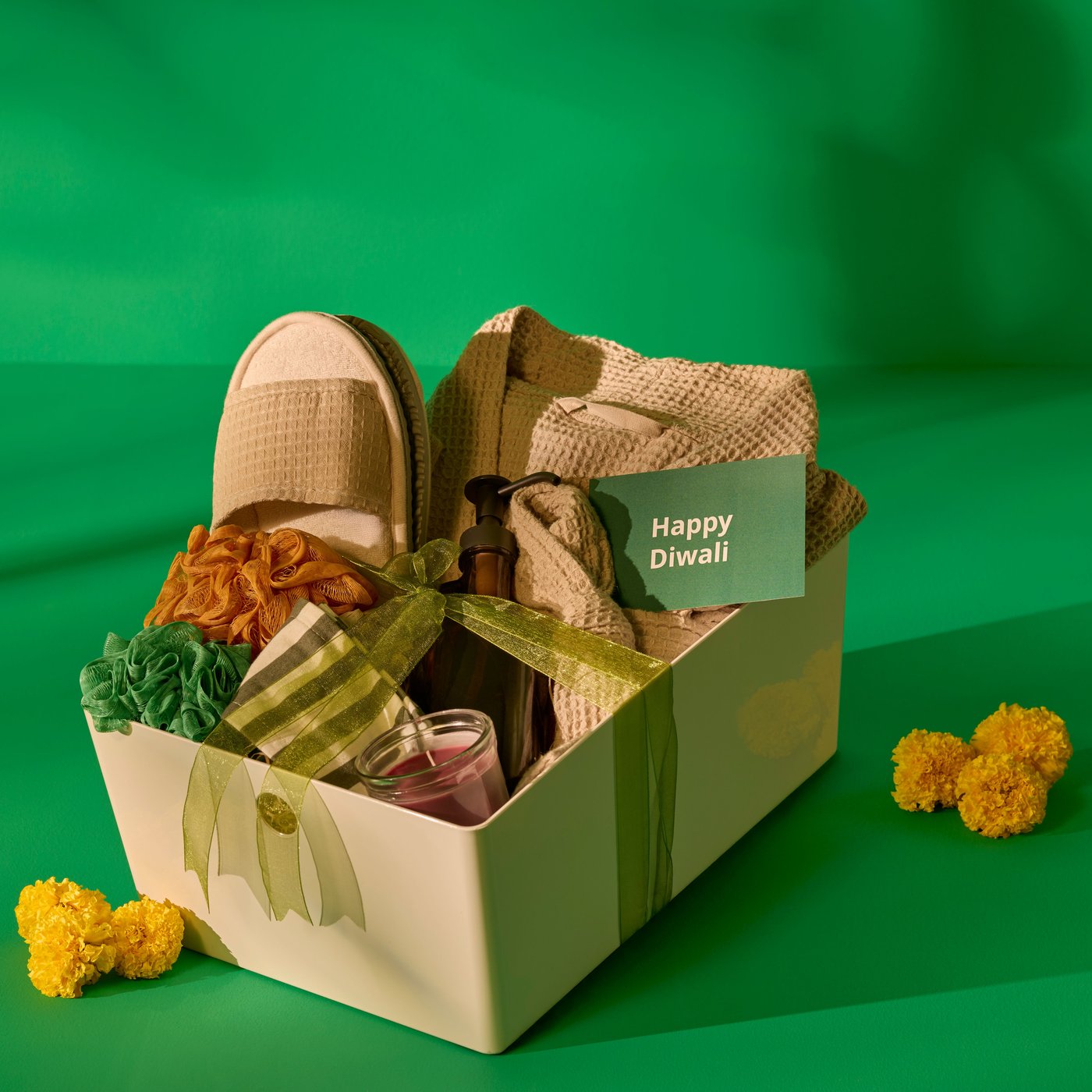 A white gift box with a bathrobe, slippers, headband, soap dispenser, scented candles, and a festive card reading 'Happy Diwali,' styled with marigold flowers against a green backdrop.