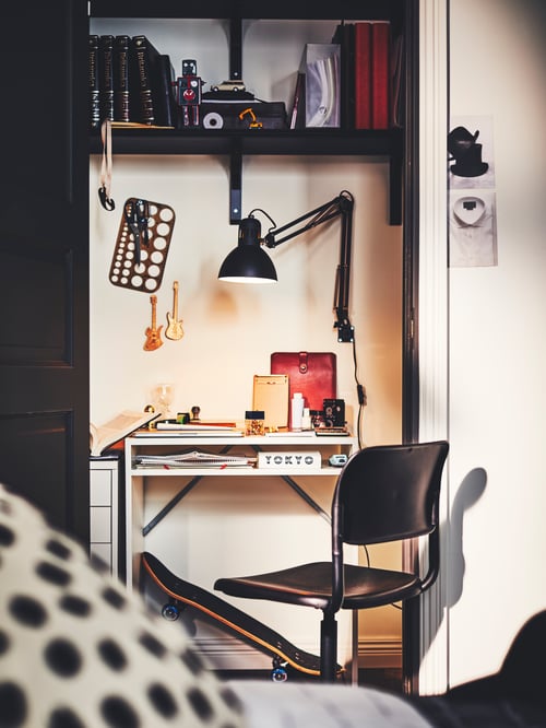 Home office with white desk, wall shelves and black plastic swivel chair with steel frame and adjustable textured seat.